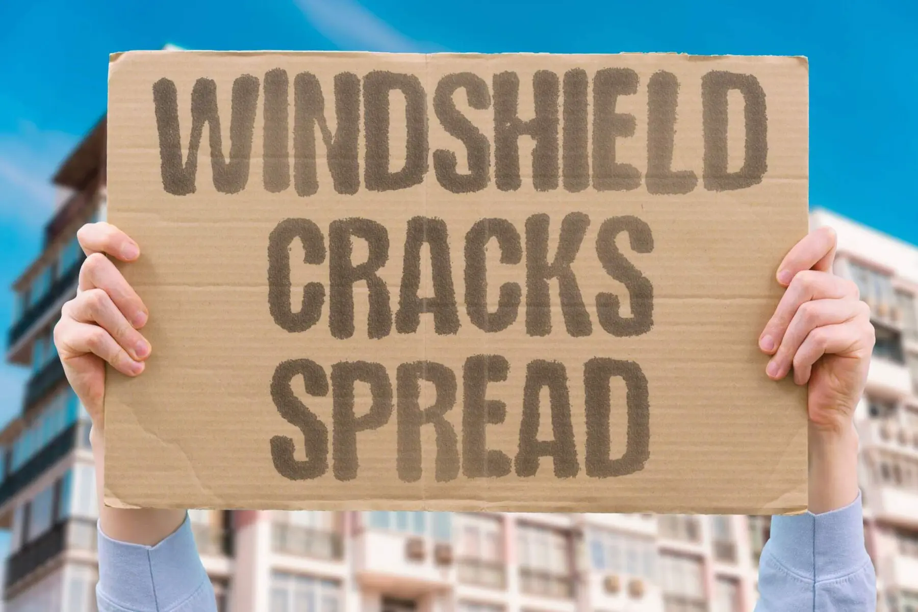 Hands holding a cardboard sign that reads Windshield Cracks Spread