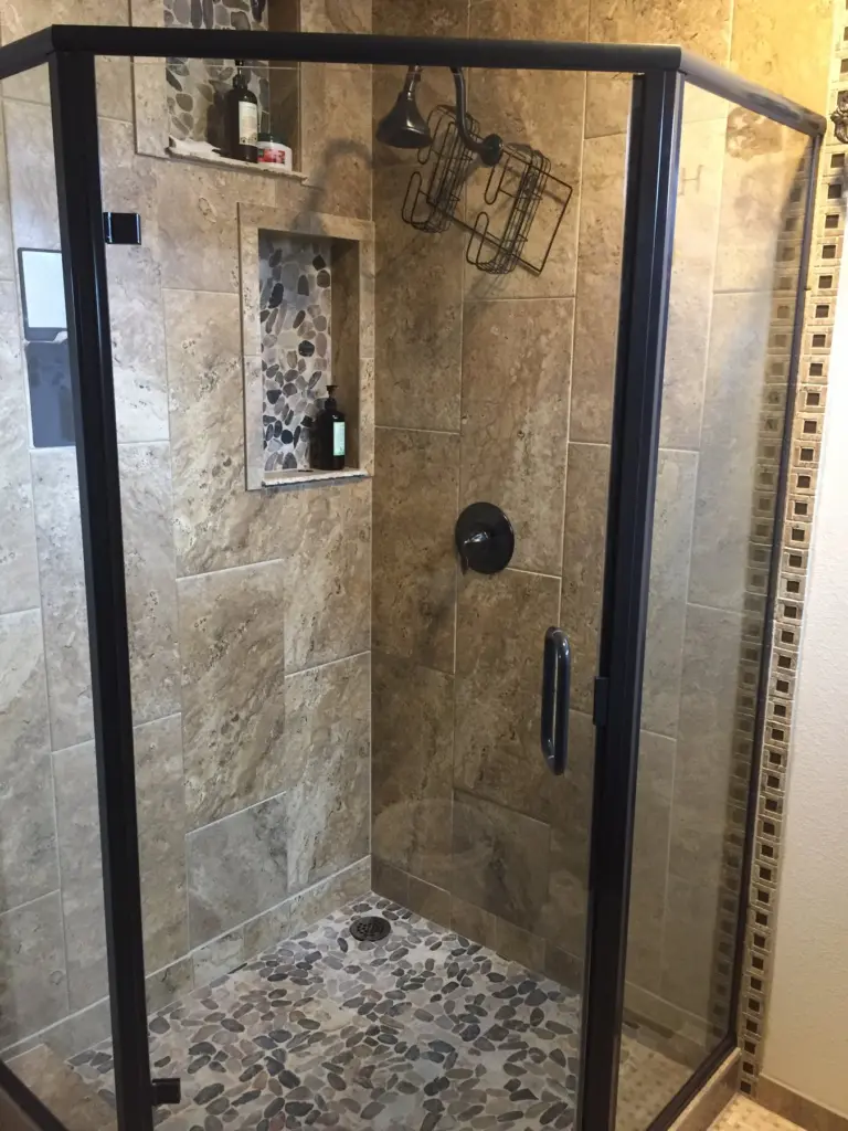 Walk-in shower with a black-framed glass enclosure, travertine tile walls and a pebble stone floor